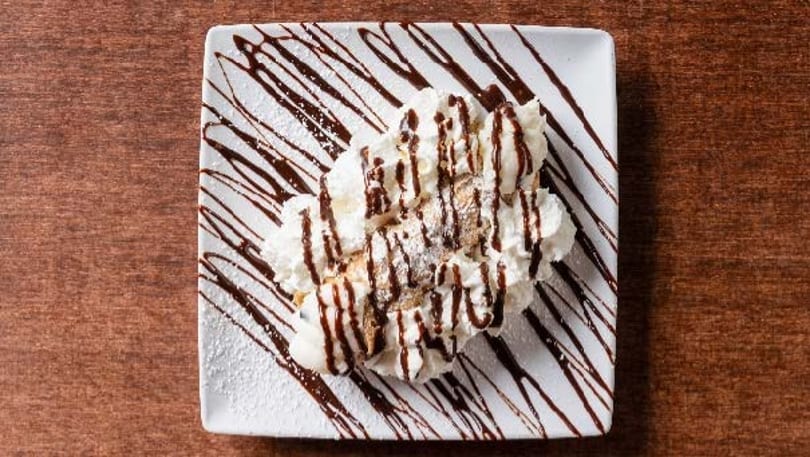 Two cannolis topped with whipped cream, chocolate syrup, and powdered sugar on a white plate.