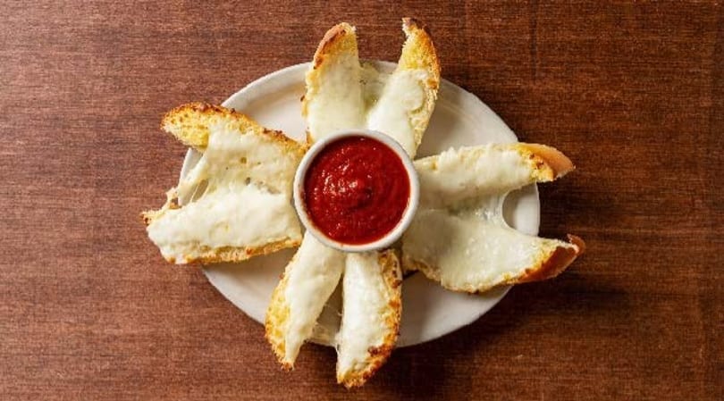 Cheesy garlic breadsticks with melted mozzarella, arranged around a small bowl of red marinara sauce.