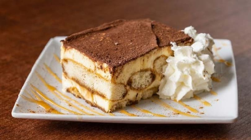 A slice of tiramisu with whipped cream and caramel drizzle on a white rectangular plate.