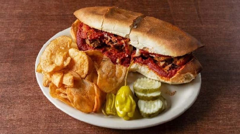 Meatball sub with potato chips, pickles, and a pepper on a white plate.